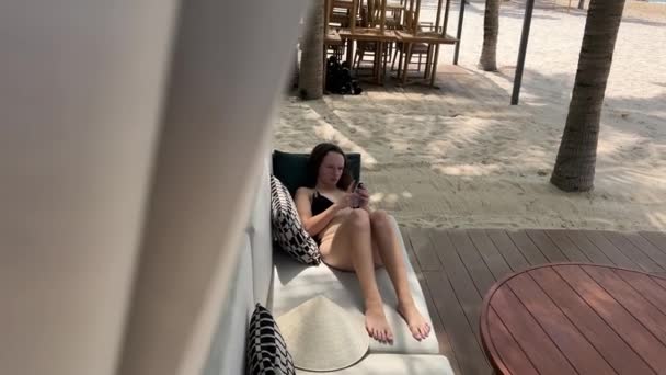 时尚奢华海滩一位身穿白色长袍的年轻女子身着黑色泳衣跳舞 躺在床上喝着水 看着手机在海面上的移动网络 Phu Quoc越南美丽的海滩 — 图库视频影像