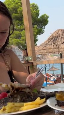 Plajdaki siyah mayo giymiş genç bir kız, Arnavutluk sahilinde Dorada balığı yiyor. Kızarmış sebzeleri beyaz tabakta kızartıyor. Öğle yemeğinde, güzel havalarda. seyahat