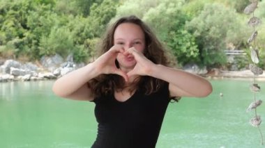 Genç Kadın 'dan el yapımı kalp işareti, nehrin yeşil rengi. Siyah elbiseli genç kız arka planda su ve yeşil ağaçlı kayalık kıyılarda poz veriyor.