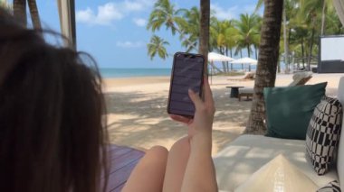 Hamakta akıllı telefon kullanan, tropikal iklimde palmiye ağaçları arasında uzanan genç bir kadın...