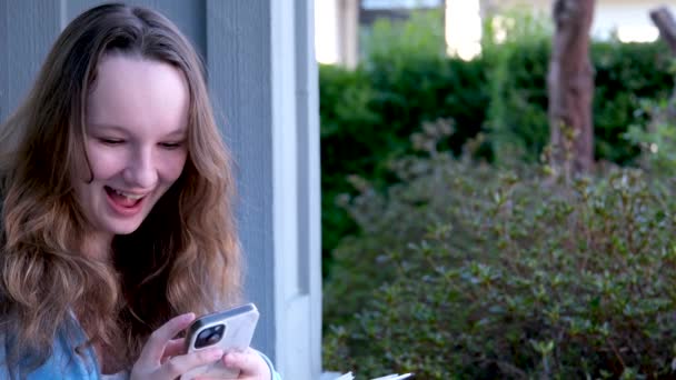 満足した幸せな十代の少女は スマートフォンでオンライン会議を話す 屋外のタッチパッドガールを選択する絵文字は 幸せな思春期の友人を笑って鼻を輝かせます — ストック動画
