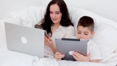 Anne ve oğlu beyaz battaniyenin altında yumuşak bir yatakta erkek tabletinin ellerinde sosyal ağlarla oynuyorlar. 4-6 yaşlarında kadın bilgisayarlı uzaktan kumanda konferansında serbest çalışıyor.
