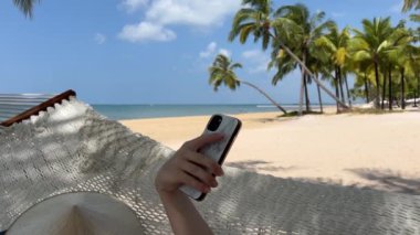 Güneş gözlüklü mutlu kadın tropik plajda palmiye ağaçlarıyla akıllı telefon kullanıyor, tatil ve yaz tatillerini sembolize ediyor. Vietnam phu Quoc adası