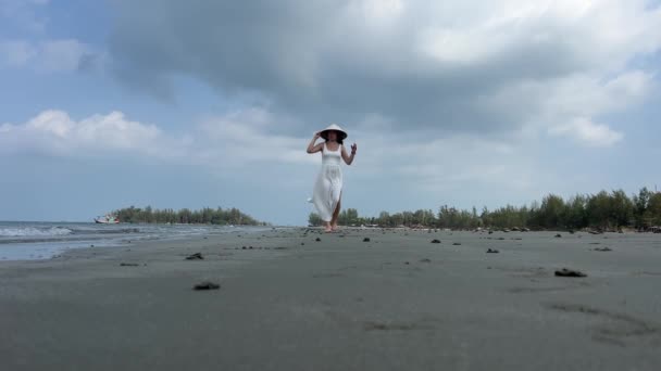 白いドレスの空のビーチにいる若い女性は 靴を履いている砂に沿って歩きます ベトナムの帽子をかぶったヨーロッパの少女 — ストック動画