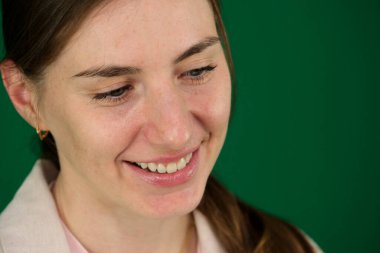 Genç sarışın beyaz kadın içeride kameraya poz verirken gülüyor. Yeşil arka planda, kromakey, bej ceketli, kırmızı manikürlü, mavi gözlü bir kadının yakın çekimi.