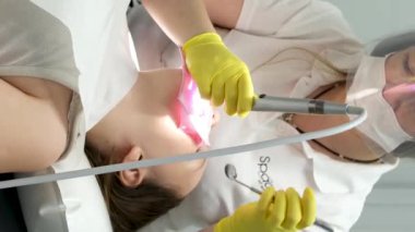 Diş hekimliği ergenliği, sarı eldivenli bir doktor dişçide dişçiye yakın çekim videosu için genç bir kızın dişini doldurur ve dişini yeni teknolojilerle kurutur. modern klinik