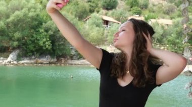 Nehrin yeşil rengi siyah elbiseli genç bir kız suyun arka planında ve ellerinde yeşil ağaçlı cep telefonuyla kayalık kıyılarda poz veriyor. Sosyal ağlar, bloglar.