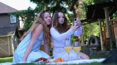 Take photos, take selfies, blog Two girls in Canada on a picnic sit on the grass on a mat, eat fruit and drink juice. Girlfriends friends LGBT relationships