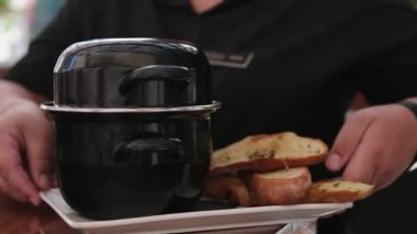 The waiter serves a black pan with croutons, croutons, delicious food, soup, serving on the table. Serving mussels
