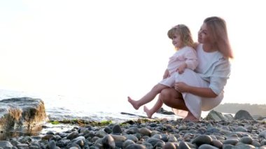 Little cute girl with mother sitting on beach earth ground playing with sand with red bucket and shovels making a castle. Woman with daughter on the beach