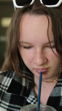 A girl drinks a drink in a cafe from a plastic glass with a straw. checkered black shirt and sunglasses.