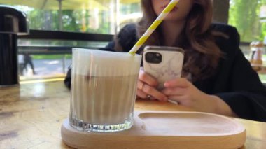 Kadınlar soğuk kahve içer ve kahve dükkanında akıllı telefon oynarlar. Buzlu latte iç. Sarı çizgili kağıt pipet içiyor.