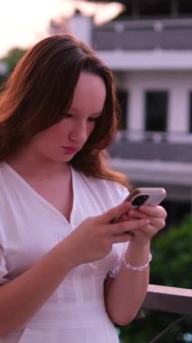 Lüks otel Avrupalı kız. Pencerenin dışındaki gece şehri temiz hava yazı. Balkonda elinde telefon olan güzel bir kadın. Beyaz resmi elbise, uzun kahverengi saç.