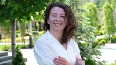 Kıvırcık saçlı, orta yaşlı kız başını çevirip kameraya bakıyor. Gülümseme flört ediyor. Eller yukarı ve baş neşe ve mutluluk