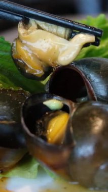 Çok fazla pişmiş salyangoz var, garsonun elinde çatal bıçak, restoranda bir misafirin tabağına yemek koyuyor. Salyangoz yemek, yemek çubuklarıyla iç çekirdeği çıkarmak. Salyangoz büfesi yemek