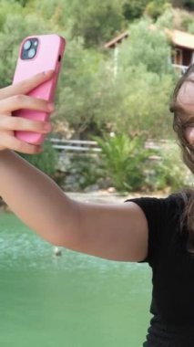 Nehrin yeşil rengi siyah elbiseli genç bir kız suyun arka planında ve ellerinde yeşil ağaçlı cep telefonuyla kayalık kıyılarda poz veriyor. Sosyal ağlar, bloglar.