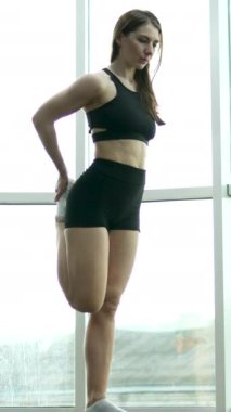 Güçlü, aktif bir kadın tek bacağıyla dengede duruyor ve antrenman odasında el ele tutuşuyor. Spor elbiseli beyaz bir kadın arka planda Fransız pencereleriyle yoga pozu veriyor..