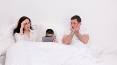 Baba ve anne gözlerini tabletten ayırmayan küçük bir çocukla yatakta oturuyorlar, sosyal ağlarda geziniyorlar, çocukluk zamanları. Gadget bağımlılığı