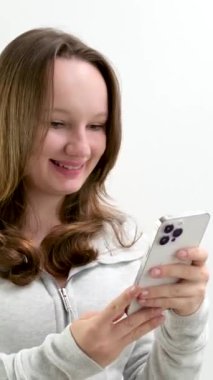 Güzel, uzun saçlı yatakta oturuyor ve akıllı telefon ekranına dokunuyor. Beyaz gömlekli, gülümseyen genç bir kadın odasında oturuyor ve telefonundaki sosyal medyayı inceliyor..