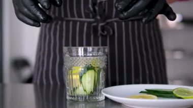 Tahta bir masanın üzerine bir bardak su, kireç ve bıçak koy. Bir adam bir bardak suya limon dilimleri atıyor. Arka planda, güneşli bir yaz gününde palmiye yaprakları ve deniz. Yavaş çekim.