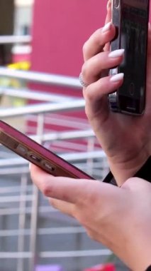 Güneş gözlüklü bir kızın elinde başka bir aletle fotoğraf çekmesi, mesaj yazması ve mesaj yazması için elinde bir telefonla iki şey yapması. Hamile kadının iki aleti var. Aynı anda iki telefon.