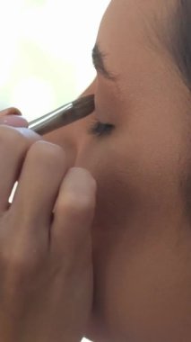 Makyajlı, tüvit ceketli, beyaz arka planda kameranın önünde göz farı sürerken poz veren şık bir kadın. Göz farını uygula