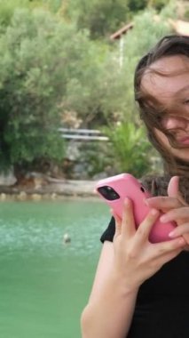 Nehrin yeşil rengi siyah elbiseli genç bir kız suyun arka planında ve ellerinde yeşil ağaçlı cep telefonuyla kayalık kıyılarda poz veriyor. Sosyal ağlar, bloglar.