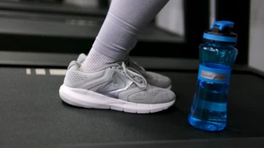 Genç, mutlu, sıska, sportif kadın, beyaz spor giyimli, ısınma trenini koşu bandının yanında su içen, pembe şişe tutan, kapalı spor salonunda başparmağını gösteren. 