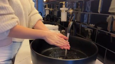 Siyah bir lavaboda ellerini yıkamak suya dokunmak ve ellerini sabunla yıkamak. Oval aynanın yanında gümüş musluk ve el kâğıdı havlularla dolu bir raf olan siyah bir lavabo. Yakın plan.