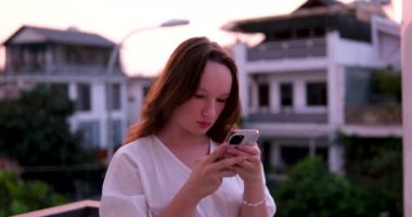 Kız akşam cep telefonu. Sokakta arka planda gece lambaları olan akıllı telefon kullanan gülümseyen kadın. Güzel bir akşamda balkonda duran beyaz elbise.