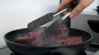 Bir şef mutfaktaki bir tavada biftek pişirir ve onu diğer tarafa çevirir, yakın plan yemek. Lezzetli biftek yapma süreci. Kızartma tavasında kızarmış et.