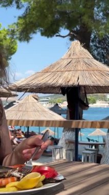 Kafkasyalı bir kız plaj restoranında tek başına yemek yiyor, omzunun arkasına bakıyor. Yalnız gezen konsept kız sahilde öğle yemeği yiyor.
