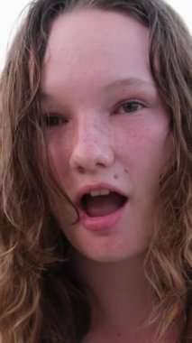 Yanmış tenli, kırmızı yanaklı, ıslak saçlı bir kızın kumsal duygularında gevşeyen hava öpücüğü. yüzme yaz tatili. Gölün arka planında bir kadın 