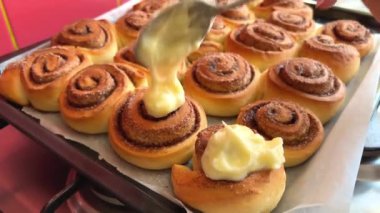 Kadın, mutfak masasındaki tarçınlı rulolara krema sürüyor. Özel spatula taze tarçınlı çöreklere beyaz tereyağı kreması sürüyor.. 