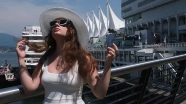 Güneş gözlüklü beyaz çerçeveli, şapkalı, beyaz elbiseli arka planda gülümseyen gözlüklü Vancouver 'daki Kanada mekanı arka planda beyaz yelken açıyor. Avrupalı kadın seyahat ediyor
