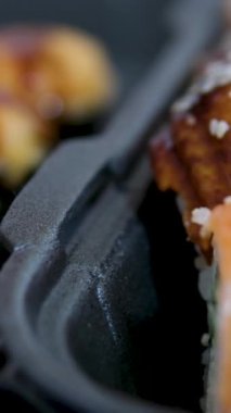 Sopalar suşi kutusundan suşi alır, yakın plan. Plastik bir kutuda yuvarlanıyor. Çeşitli lezzetli Japon suşilerinden oluşan hazır yemek seti teslimat konteynerinde. Sağlıklı beslenme, Asya yemekleri, hazır yemek.