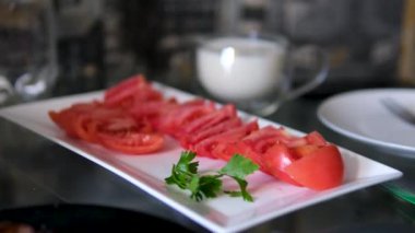 Taze dilimlenmiş domatesler maşayla beyaz bir tabakta, sıcak ışıkla aydınlatılmış mutfak ortamında servis edilmeye hazır. Beyaz tabakta domates dilimleri