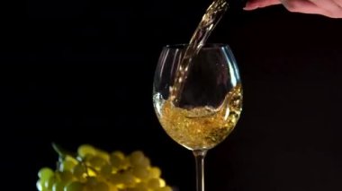 Siyah arka planda beyaz şarap yeşil üzümlerin yanında şişeden cama şarap dökülüyor. Kuru üzüm.