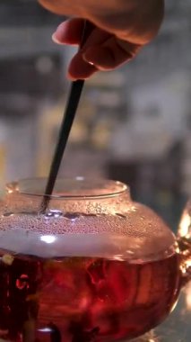 Sudan gülü çiçeklerinden cam çaydanlıkta kırmızı amber çayı yapma süreci. Ve mum ışığında koyu bir arka planda cam çaydanlıkta bir kaşıkla kırmızı amber çayı karıştır.