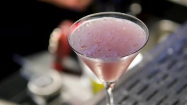 Barmen, martini bardağında pembe bir kokteyl süslüyor. Siyah eldivenler giyiyor. İçki sunumunda profesyonelliği ve zarafeti vurguluyor.