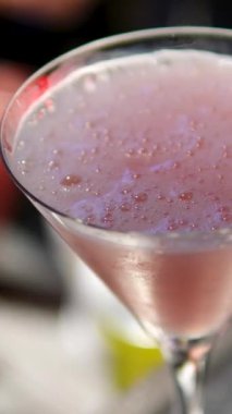 Barmen, martini bardağında pembe bir kokteyl süslüyor. Siyah eldivenler giyiyor. İçki sunumunda profesyonelliği ve zarafeti vurguluyor.
