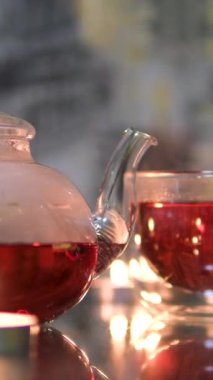 Bir fincan kırmızı çay ve cam çaydanlık, altında bir mum ile ısıtılan bir stantta oturur. Bir yudum almak için bir fincan kaldırılır. Bir kadının eli masadaki mumları ayarlar.