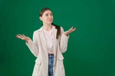 Yeşil arka planda, kromakey, bej ceketli, kırmızı manikürlü, mavi gözlü, stüdyoda kafası karışmış bir kadının portresi.