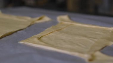 Elleri ahşap yüzeyde yuvarlanan hamurun yanında merdane ve un var. Un kaplı masada yuvarlanan aletle çalışırken parmaklar ince hamur çarşaflarını şekillendiriyor. Eller hamuru düzleştiriyor. 