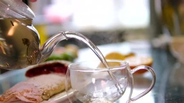 Mutfak masasında sıcak çay yapıyorum. Kaynar su çaydanlıktan bir çay poşeti ile bir fincana dökülür. Güneşli bir günde mutfak masasında şeffaf taze demlenmiş çay. Yüksek kalite 4k