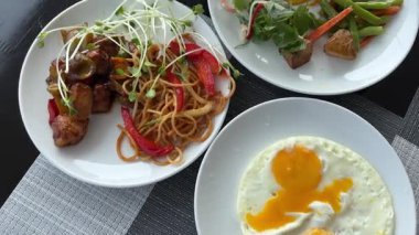 Öğle yemeğinde kızarmış yumurta, beyaz tabak makaronlu spagetti, mikroyeşillik ve et sebzeli tavuk, siyah bir masada çiçekler ve peçeteli şık bir sunum. lüks 