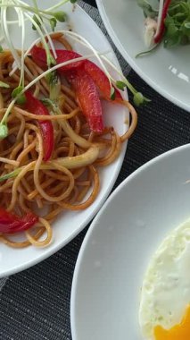 Öğle yemeğinde kızarmış yumurta, beyaz tabak makaronlu spagetti, mikroyeşillik ve et sebzeli tavuk, siyah bir masada çiçekler ve peçeteli şık bir sunum. lüks 