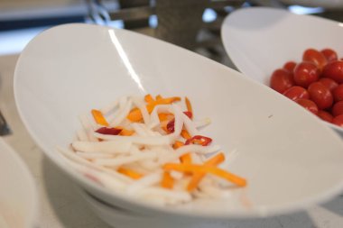 Soyulmuş taze havuç ve daikon turpu dilimleri, soyulmamış daikon turplarının yarısı beyaz bir masada yatıyor. Havuç, Daikon turpu, sebze salatası ve abur cuburlarda popüler malzemeler.