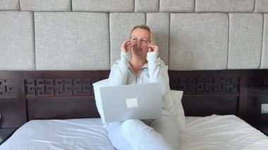 Yetişkin bir kadın dizüstü bilgisayarıyla yatakta oturuyor, gözlüklerini çıkarıyor göz dinlendirici göz egzersizleri yapıyor, işini bitiriyor sosyal ağlarda internet gezici iletişimiyle yatakta hayatın tadını çıkarıyor. Yüksek kalite 4k 