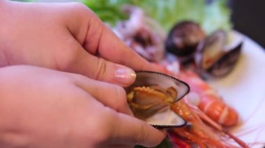 El, buğulanmış deniz tarağı kabuğundan eti açar ve çıkarır. İçindeki taze eti ortaya çıkarır. Arka planda toplanan bir kase midye eti de deniz ürünlerinin hazırlanma anını betimler..